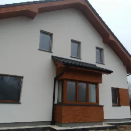 Wykończony dom jednorodzinny z jasną elewacją, drewnianymi elementami wykończeniowymi wokół okien i pod dachem, oraz zadaszeniem nad oknem balkonowym, widoczne małe okna na poddaszu.
