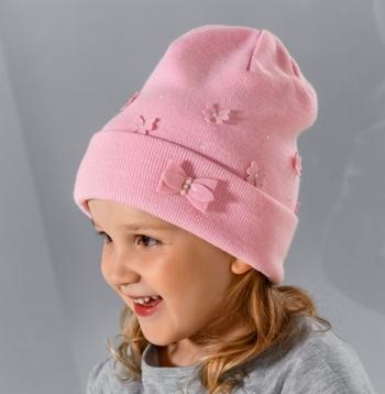 Uśmiechnięta dziewczynka w różowej, dzierganej czapce z aplikacjami w kształcie kwiatków i kokardek, na szarym tle.