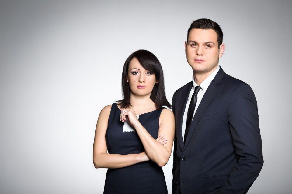 Portret dwojga prawników: kobieta w granatowej sukience z identyfikatorem i mężczyzna w ciemnym garniturze na szarym tle.