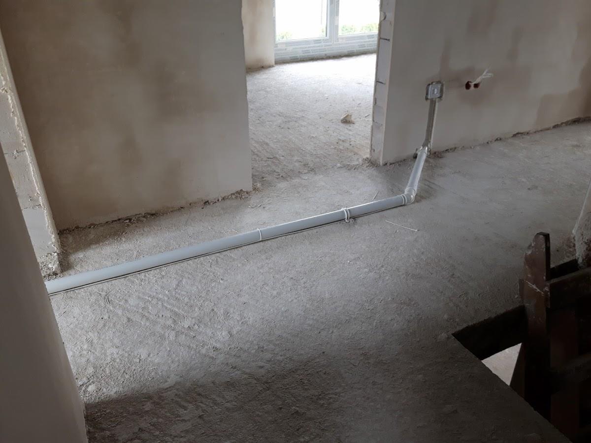 Szara rura instalacyjna prowadzona po podłodze w surowym wnętrzu, z widoczną puszką elektryczną na ścianie i wystającymi przewodami, w tle okno i widok na inne pomieszczenie w stanie deweloperskim.