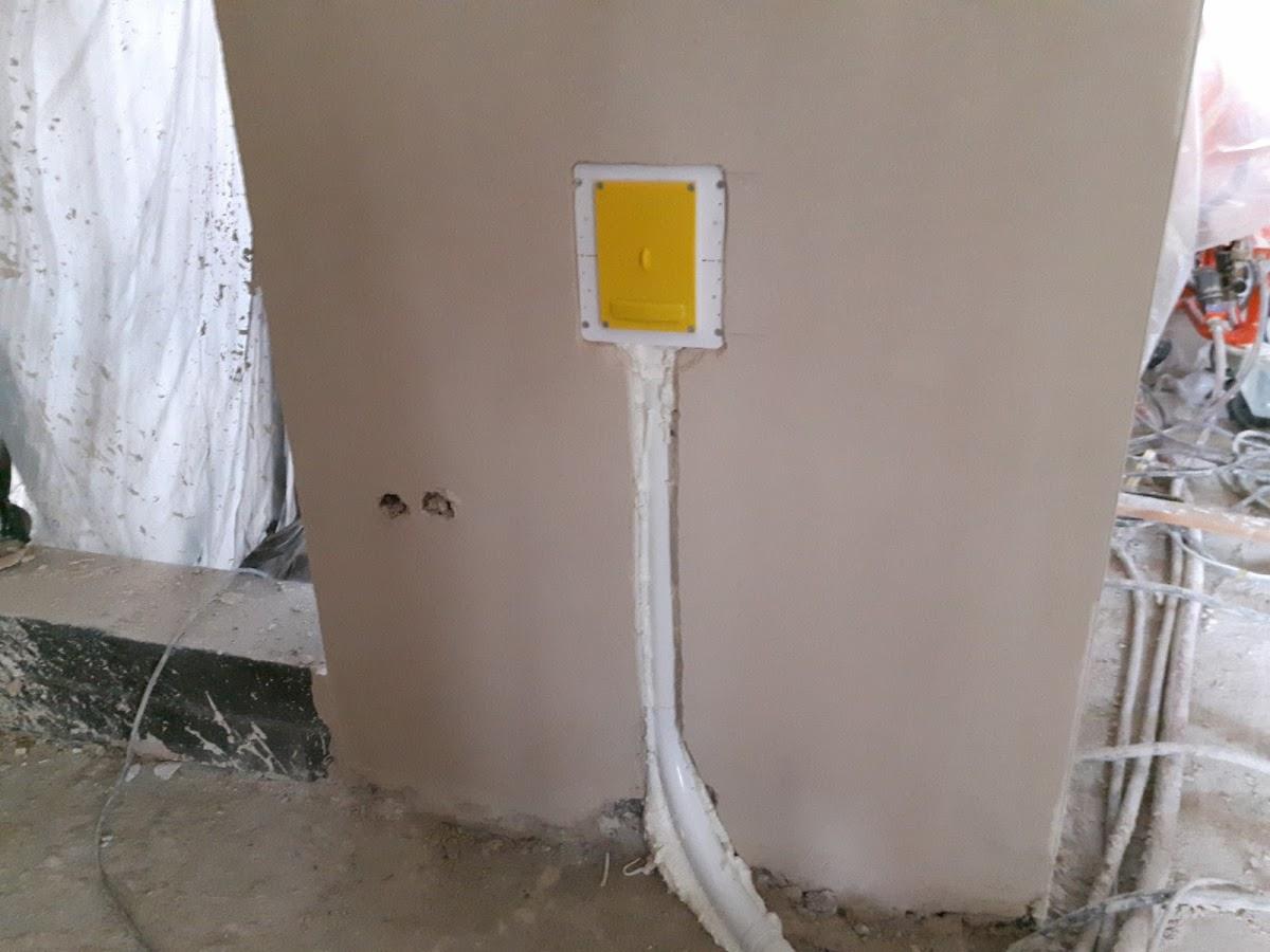 Żółta skrzynka elektryczna w ścianie, białe rurki z kablami zabezpieczone pianką montażową, widoczne dwa otwory na kable w ścianie, w tle bałagan budowlany.