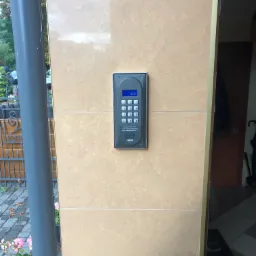 Szary domofon cyfrowy ACO zamontowany na beżowej, kafelkowanej ścianie przy wejściu do budynku, widok z zewnątrz, z fragmentem słupka ogrodzenia i roślinami w tle.