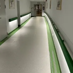 Długi, jasny korytarz w budynku użyteczności publicznej, z zielonymi akcentami kolorystycznymi na ścianach i podłodze, oświetlony lampami LED. Na ścianach wiszą obrazy w ramkach.