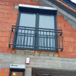 Szara, metalowa balustrada okienna zamontowana na elewacji budynku w trakcie budowy, widoczne czerwone pustaki i szara zaprawa, okno z roletą zewnętrzną.