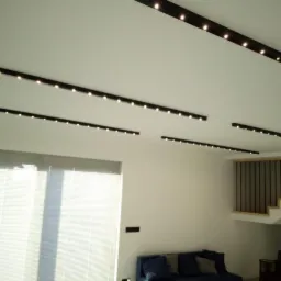 Nowoczesne oświetlenie sufitowe z czarnymi listwami i punktowymi źródłami światła, widok na schody z metalowymi balustradami i częściowo zasłonięte okno z żaluzjami.