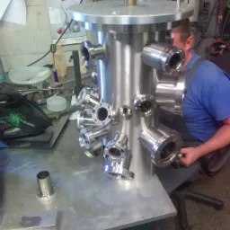 Spawacz przy pracy nad dużą, cylindryczną konstrukcją ze stali nierdzewnej z wieloma przyłączami kołnierzowymi, widoczne narzędzia i wyposażenie warsztatowe w tle.