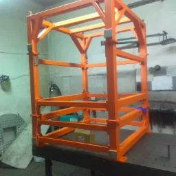 Pomarańczowa, metalowa konstrukcja szkieletowa z poprzeczkami, z widocznymi spawami, stojąca na perforowanym stole warsztatowym w hali produkcyjnej.