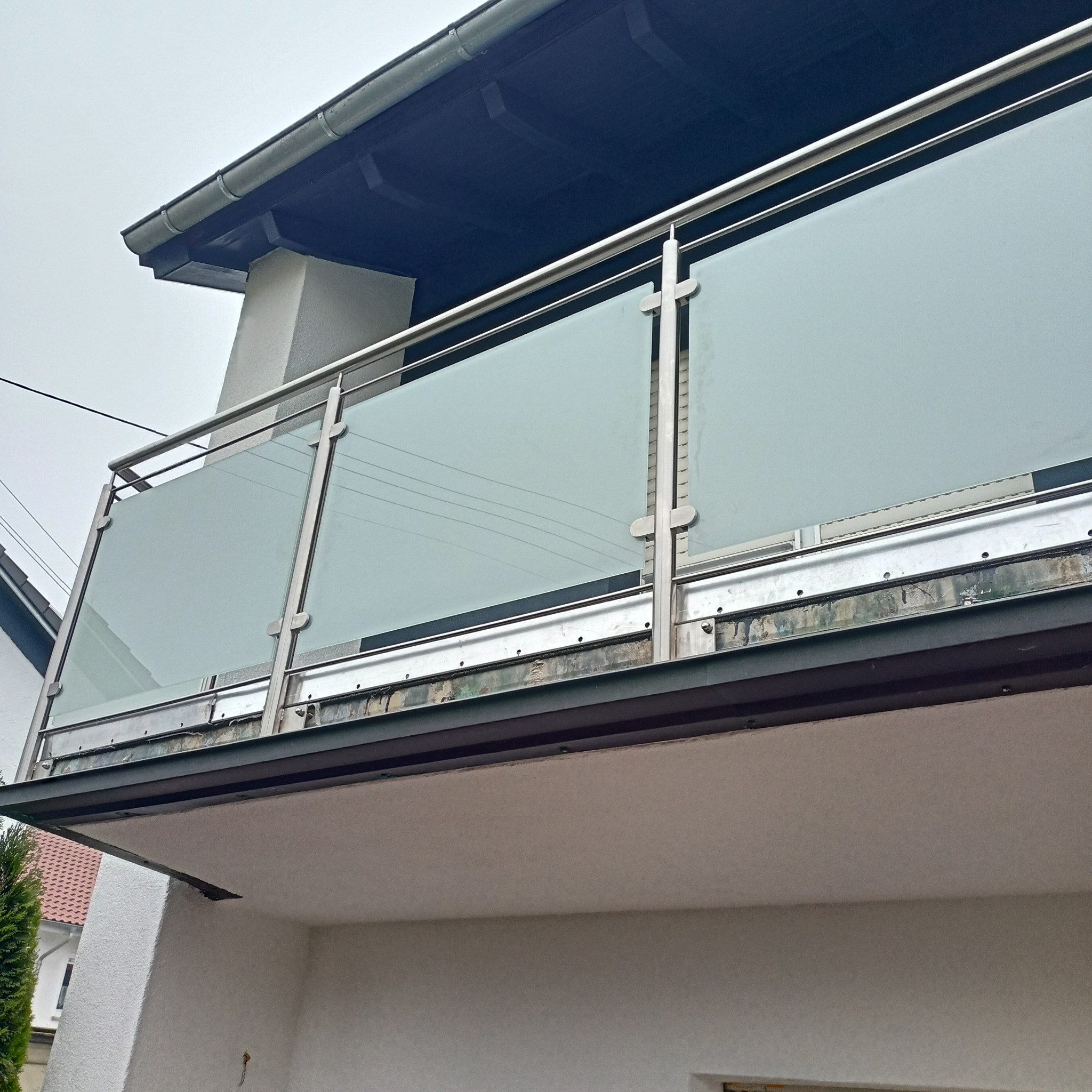 Balkon z balustradą ze stali nierdzewnej i matowego szkła, widoczne mocowania szkła do słupków, ujęcie od dołu.