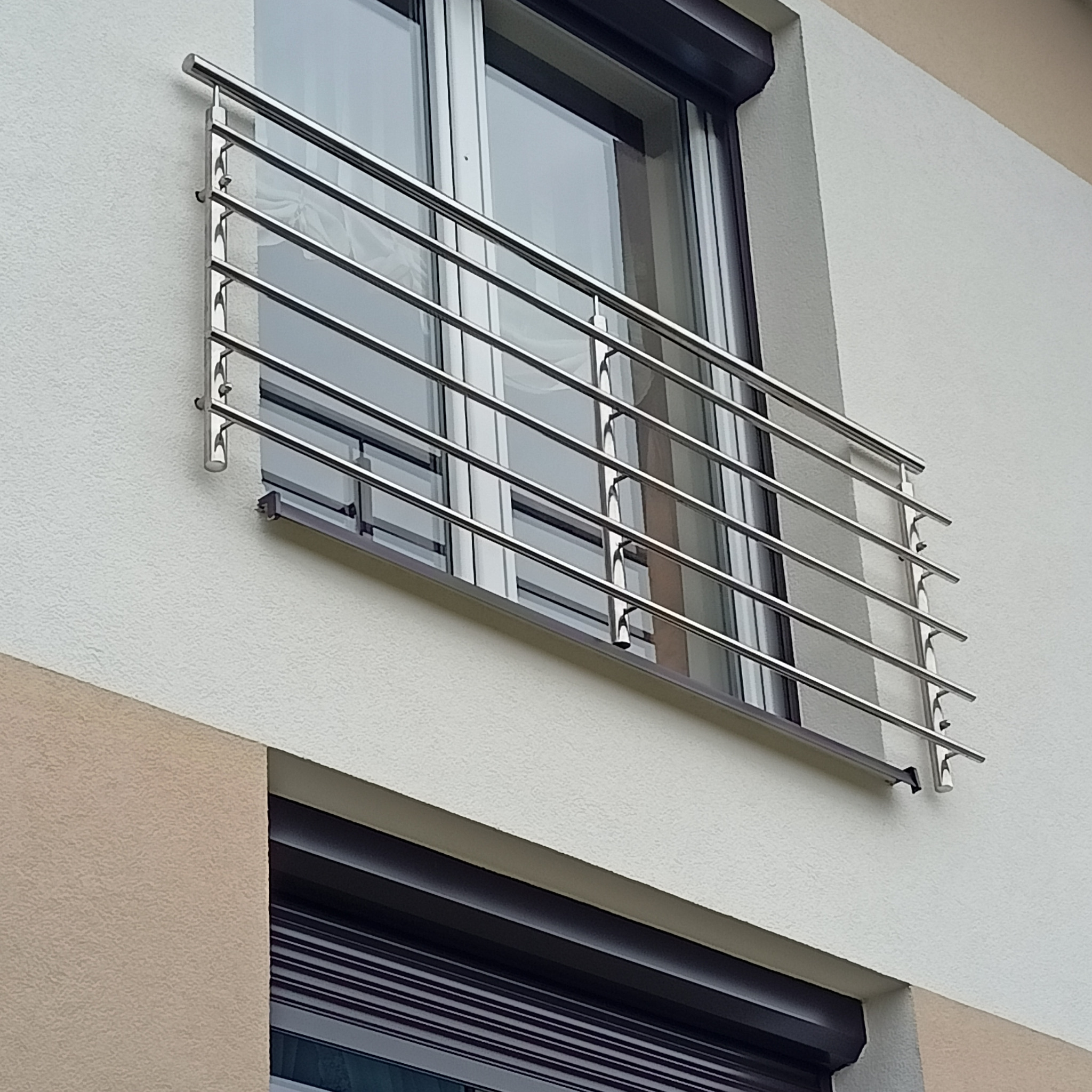 Nierdzewna, pozioma balustrada balkonowa z rurek, zamontowana na elewacji budynku z oknem i brązową roletą zewnętrzną.