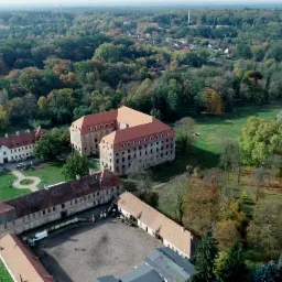 Widok z lotu ptaka na rozległy kompleks pałacowy z czerwonymi dachami, otoczony parkiem i lasem w jesiennej kolorystyce, z widoczną wieżą kościelną w prawym dolnym rogu.