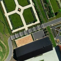 Widok z lotu ptaka na starannie zaprojektowany ogród z geometrycznymi rabatami, ścieżkami i budynkami z dachami o różnej kolorystyce, ukazujący dbałość o detale i kompozycję.