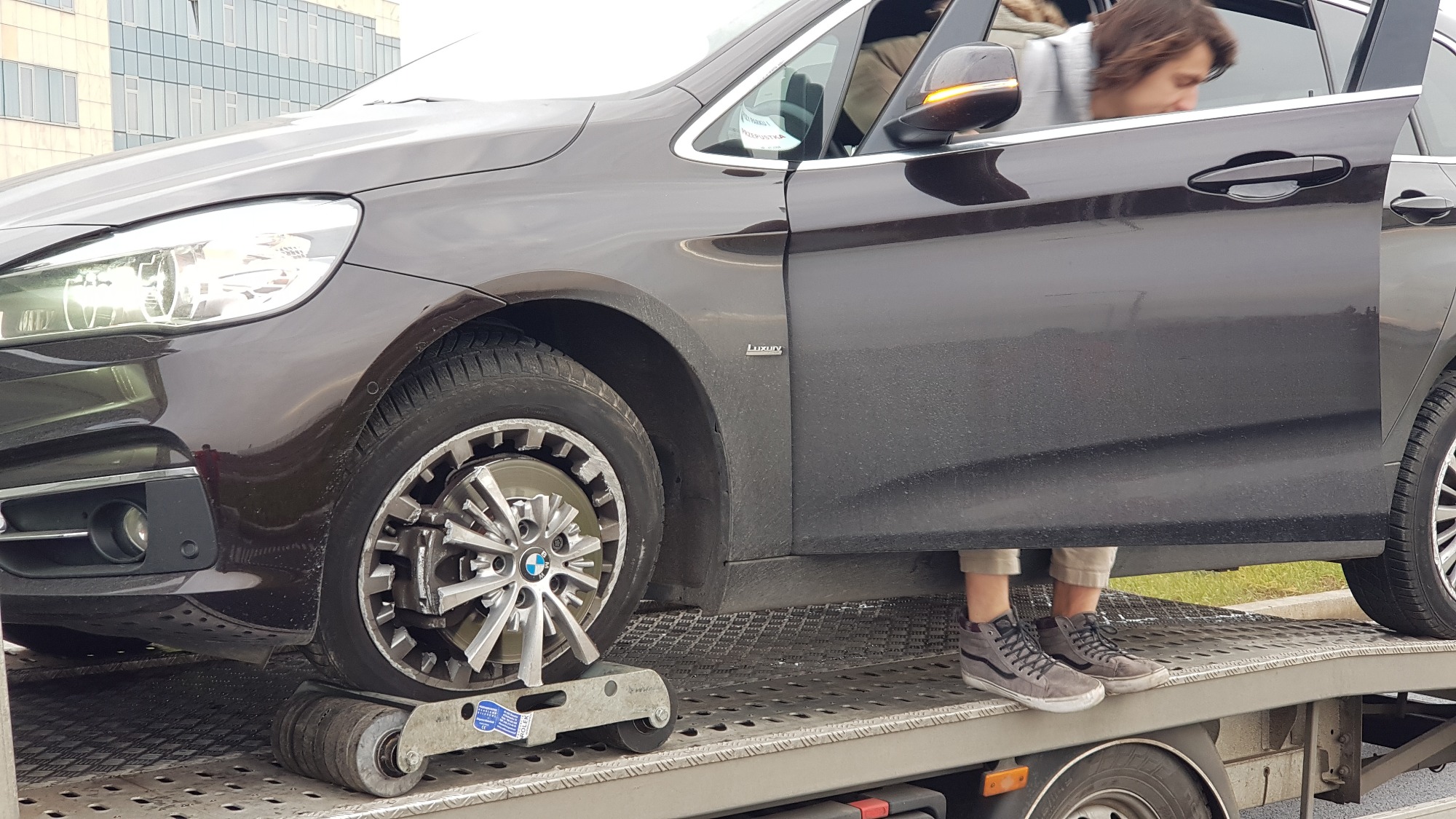Brązowy samochód BMW na platformie lawety z otwartymi drzwiami, koło zabezpieczone rolkami transportowymi, widoczne nogi osoby w szarych butach.