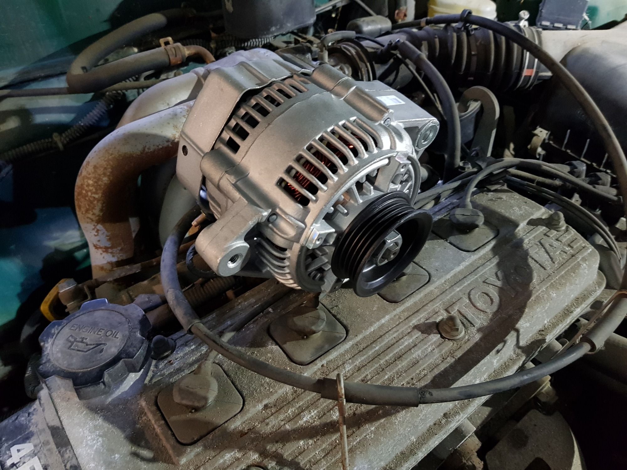 Nowy alternator na silniku Toyota, widoczny korek wlewu oleju, ogólny stan silnika wskazuje na starszy model pojazdu.