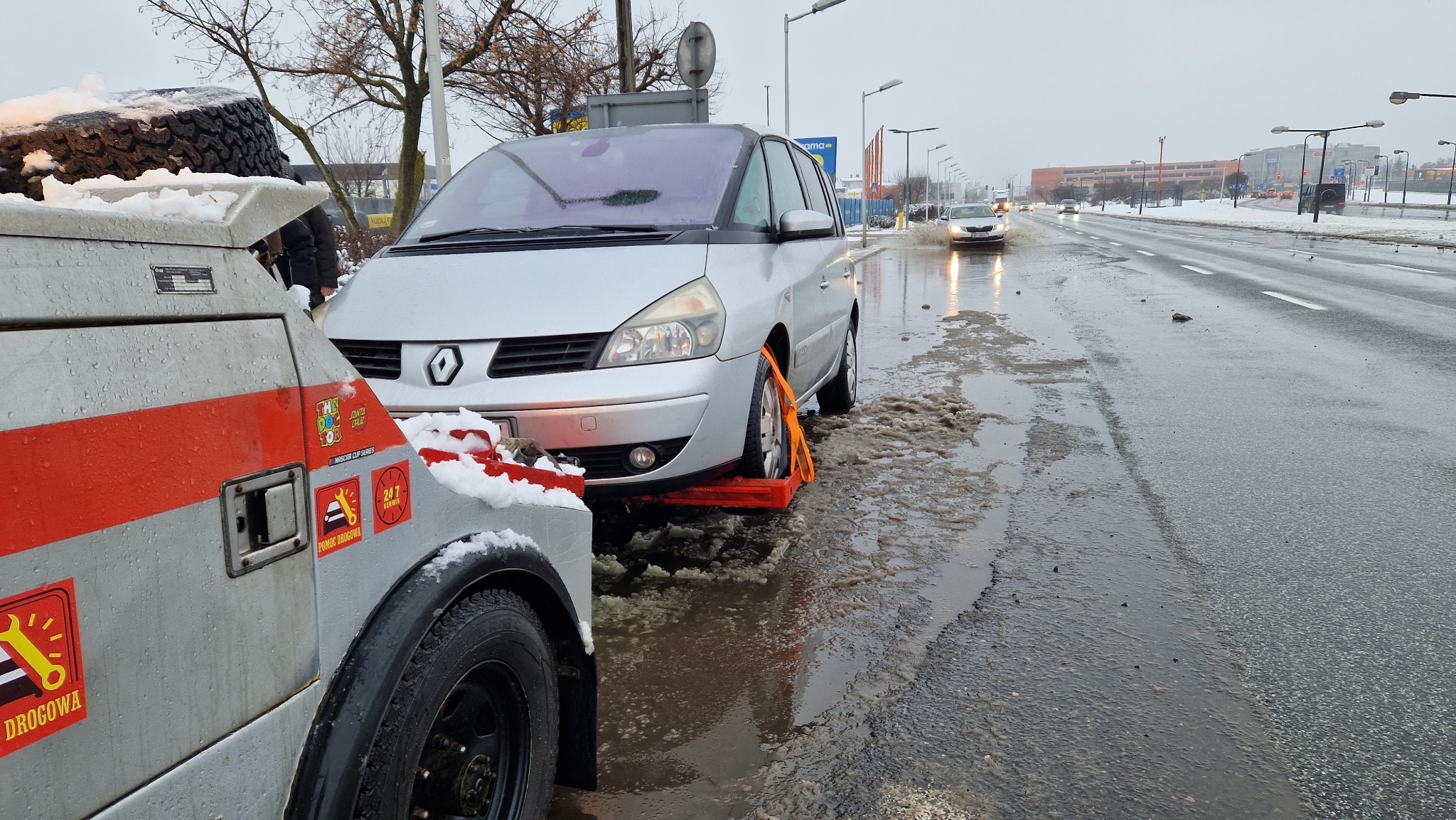 Srebrny Renault Espace na lawecie w zimowych warunkach, widoczny fragment lawety z naklejkami 'Pomoc Drogowa' i '24/7 Serwis', mokra jezdnia z resztkami śniegu.