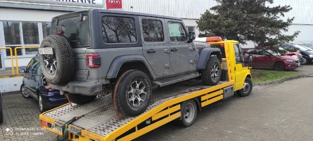 Szary Jeep Wrangler na żółtej lawecie, na tle salonu samochodowego z logo KIA.