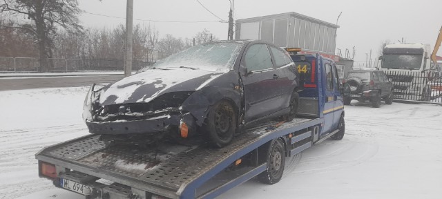 Uszkodzony, granatowy samochód osobowy na platformie lawety w zimowych warunkach. Widoczne zniszczenia przodu pojazdu i śnieg na karoserii.