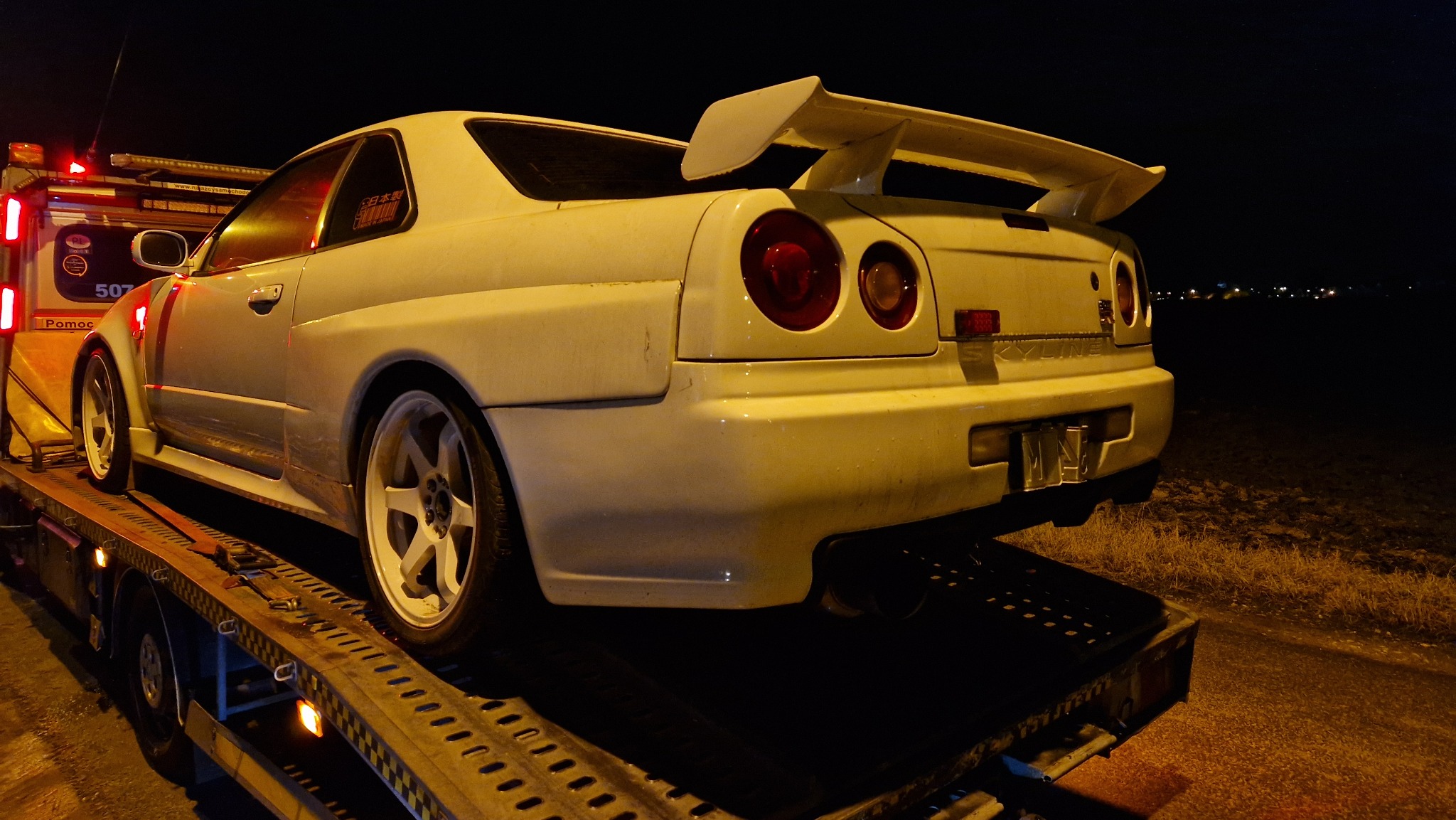 Biały sportowy samochód Nissan Skyline R34 na platformie lawety, widok z tyłu w nocnej scenerii z widocznymi światłami miasta w oddali.