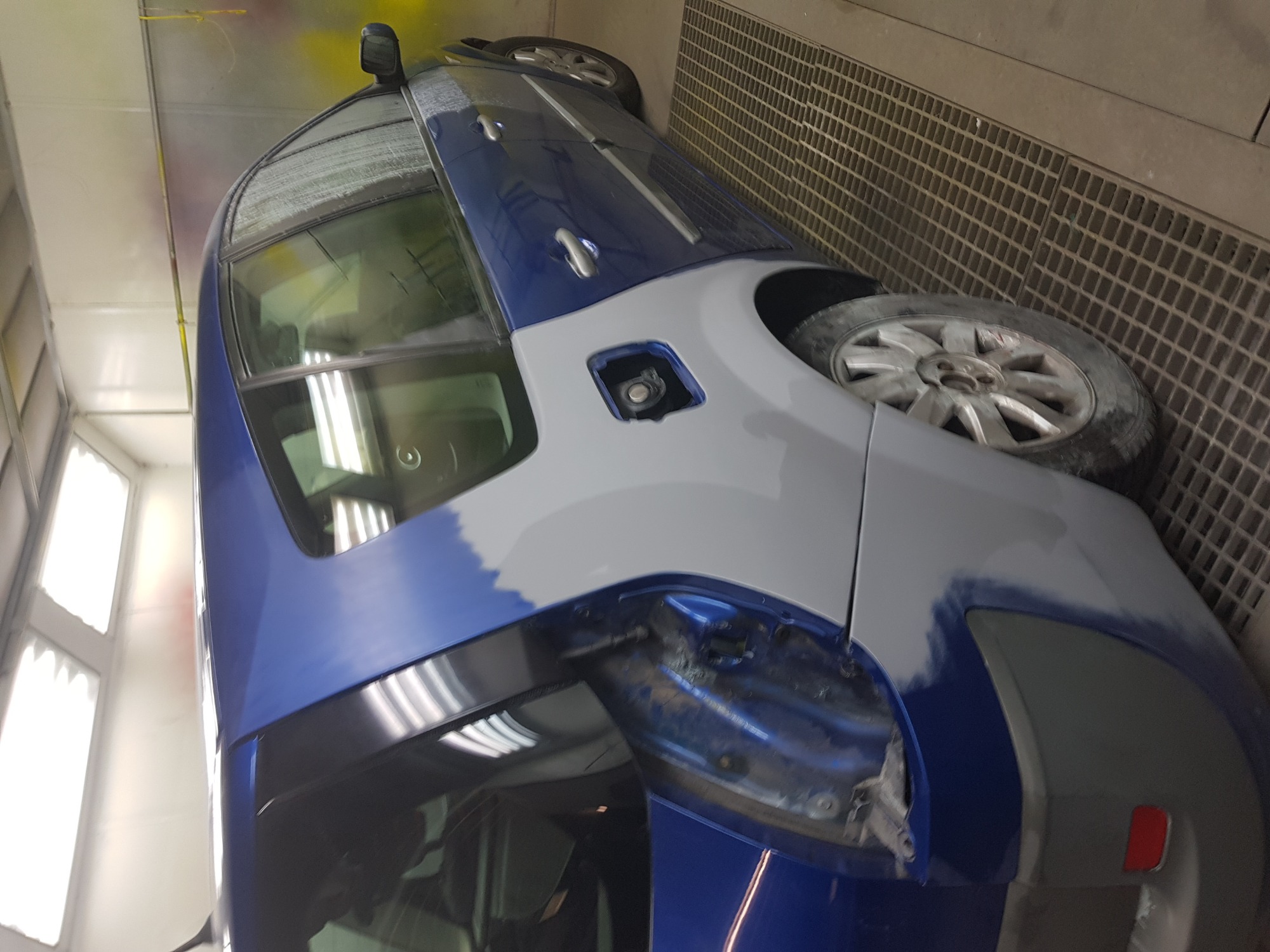 Niebieski samochód osobowy w trakcie naprawy blacharskiej i lakierniczej w warsztacie, z widocznymi śladami szpachli i podkładu na karoserii.