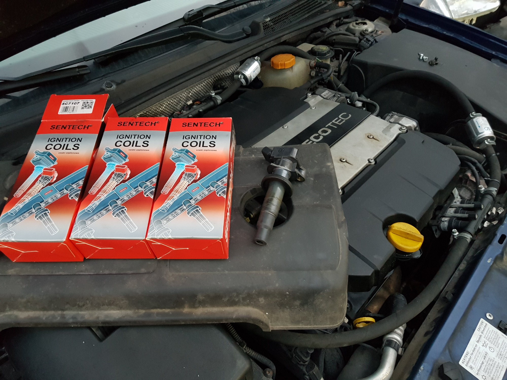 Otwarta maska samochodu z widocznym silnikiem, na którym leżą trzy pudełka z cewkami zapłonowymi Sentech SC7107 oraz jedna cewka zapłonowa obok pudełek.
