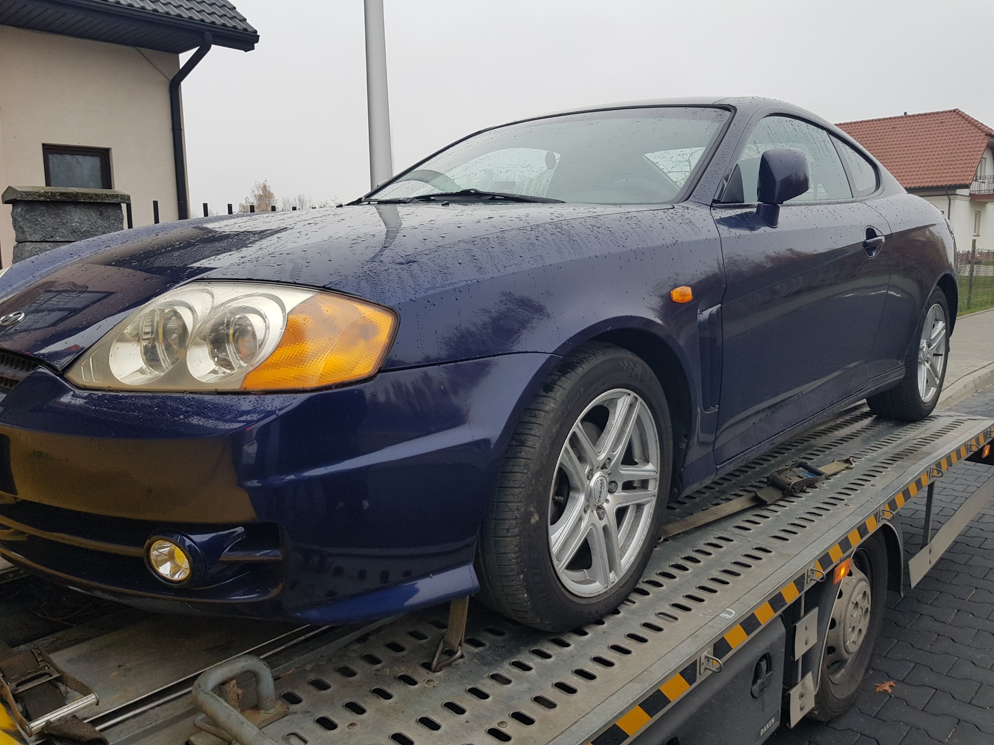 Granatowy samochód sportowy na platformie lawety, zabezpieczony pasami transportowymi, z widocznymi kroplami deszczu na karoserii.