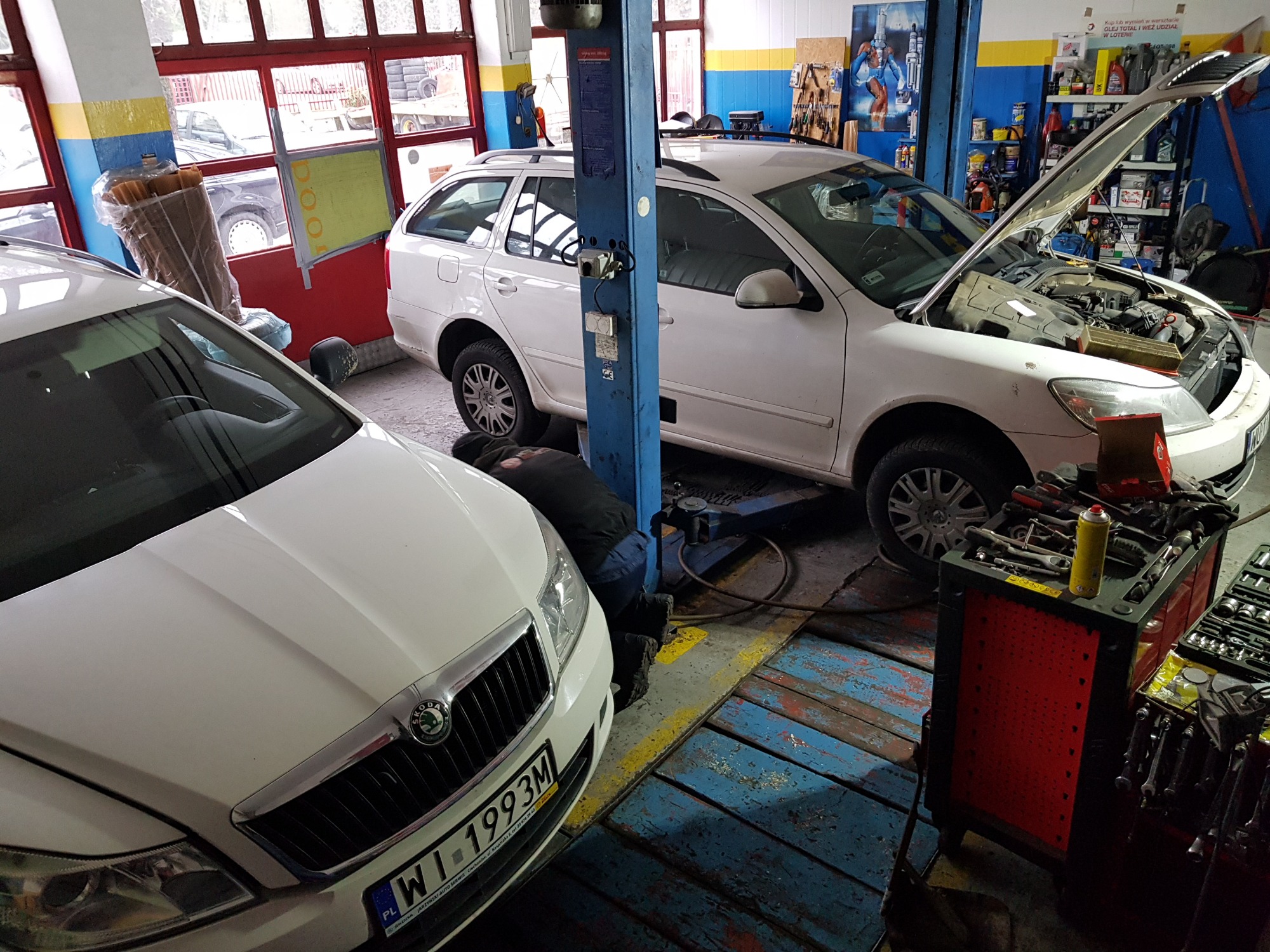 Mechanik w warsztacie samochodowym pracuje pod podniesionym białym kombi Skoda Octavia, narzędzia na wózku obok, widoczny numer rejestracyjny pojazdu z przodu.