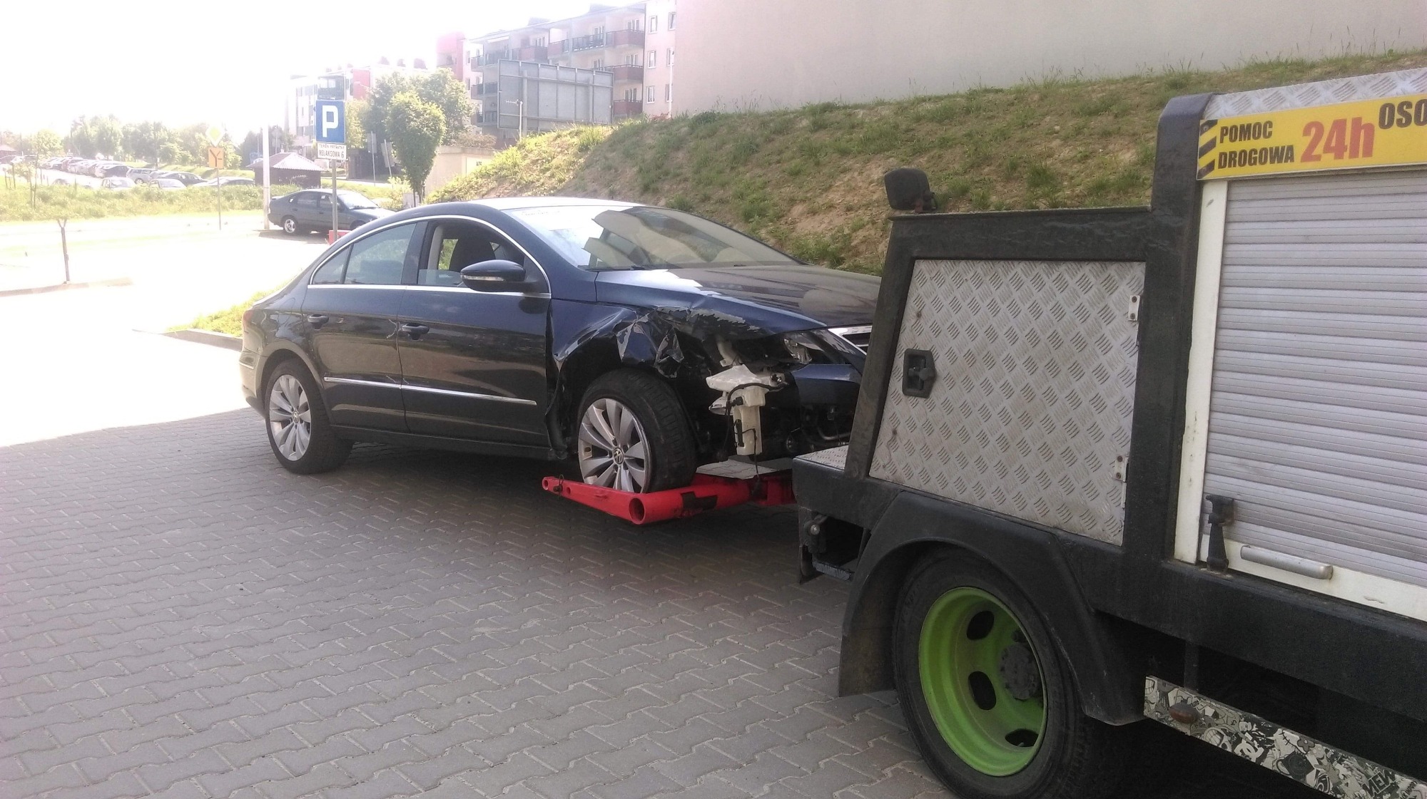 Uszkodzony, ciemny samochód osobowy marki Volkswagen Passat CC z widocznymi uszkodzeniami przodu, załadowany na czerwoną platformę lawety, z lawetą z zielonymi felgami na tle chodnika i budynku...