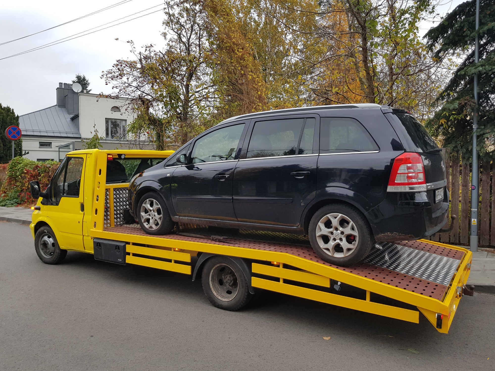 Żółta laweta z czarnym samochodem osobowym Opel Zafira na platformie, zaparkowana na ulicy w jesiennej scenerii.