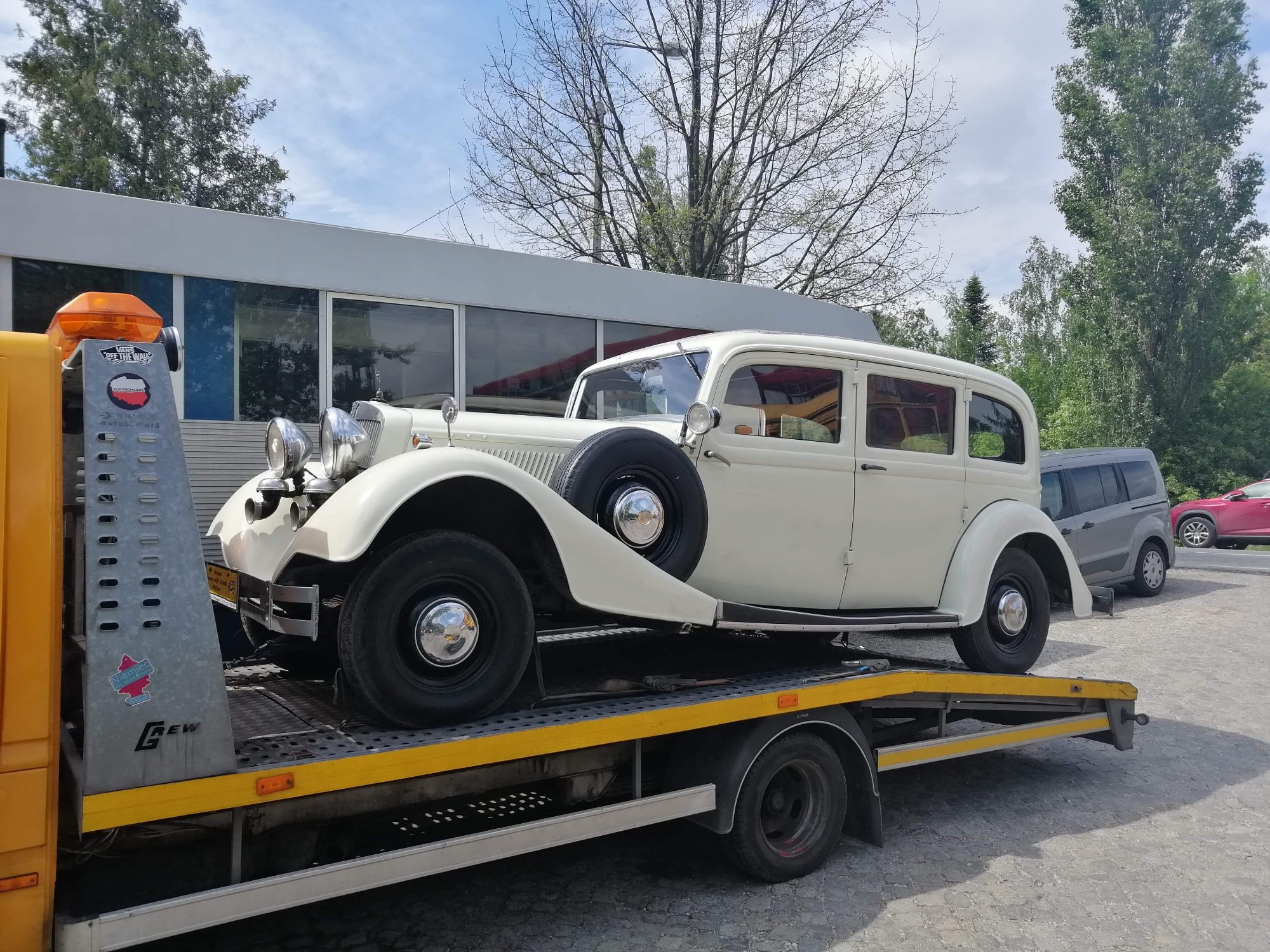 Klasyczny, kremowy samochód osobowy z kołem zapasowym na błotniku, transportowany na żółtej lawecie z włączonym pomarańczowym światłem ostrzegawczym.