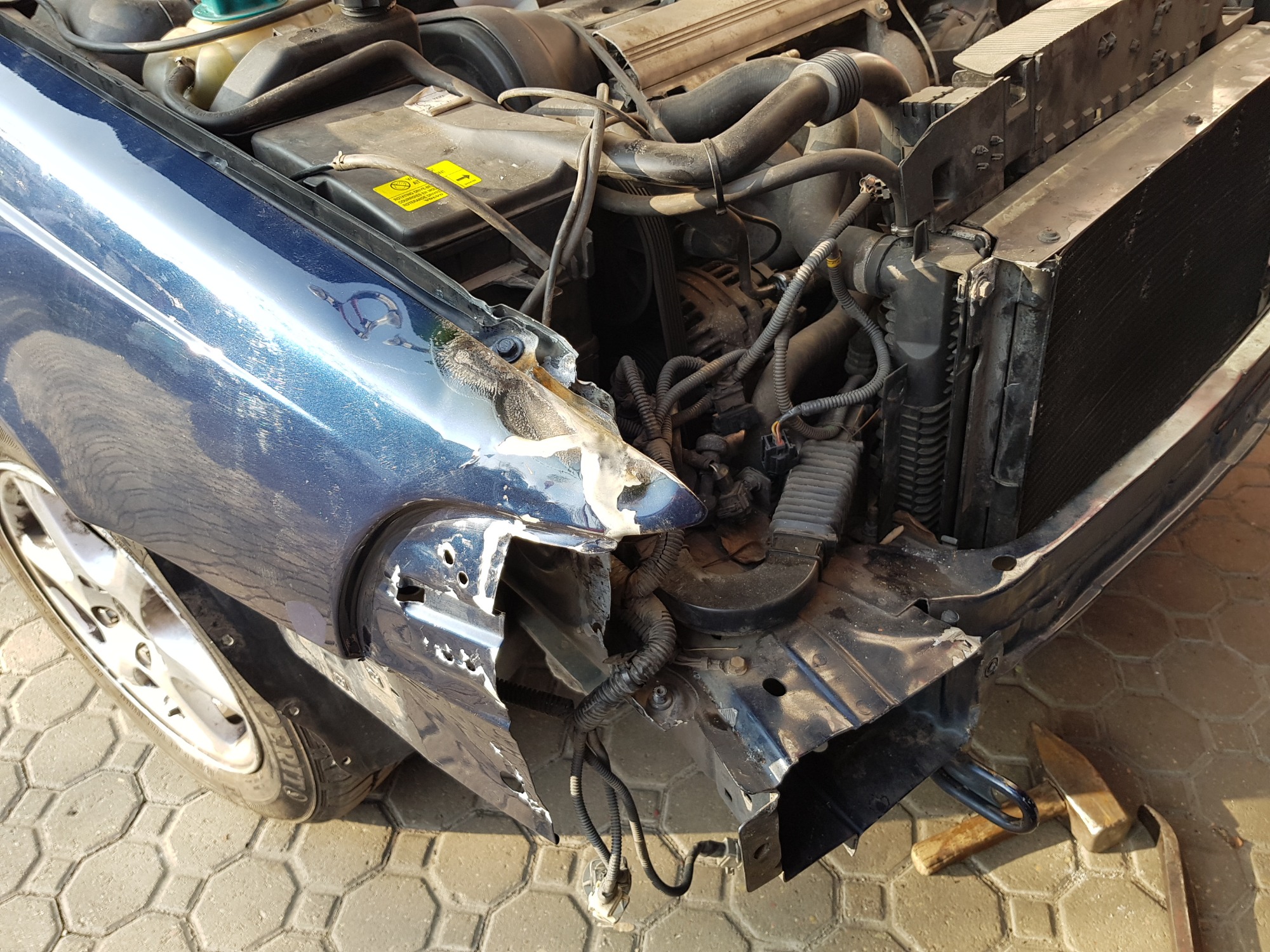 Uszkodzony przód niebieskiego samochodu z widocznym silnikiem, chłodnicą i elementami karoserii przed naprawą. Na ziemi leżą narzędzia.