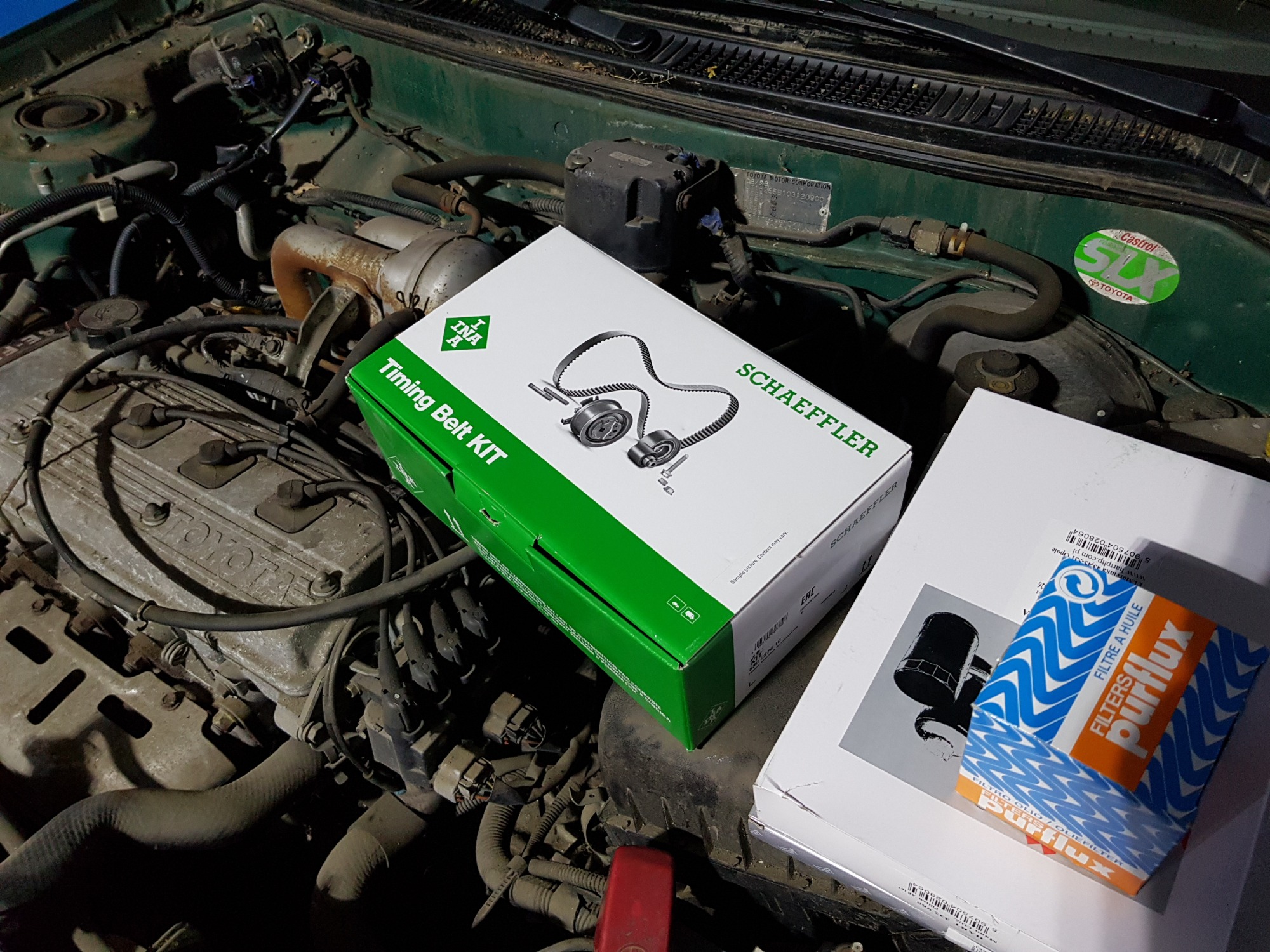 Kompletny zestaw rozrządu INA Schaeffler i filtr oleju Purflux na tle brudnego silnika Toyota, widoczna naklejka Castrol SLX. Przygotowanie do wymiany części samochodowych.