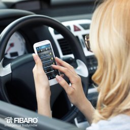 BAJER Bartosz Bajerlein - Kobieta w aucie steruje oświetleniem domu za pomocą aplikacji na smartfonie. Widoczny interfejs aplikacji Fibaro. Inteligentny dom.