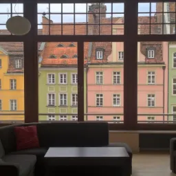 Salon z dużym oknem z widokiem na kolorowe kamienice, ciemnoszara sofa z czerwoną poduszką i fotel, parkiet na podłodze.