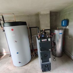 HEGAM - Instalacja kotła na paliwo stałe Hegam z zasobnikiem Emet w surowym pomieszczeniu. Widoczne rury miedziane i elementy instalacji.