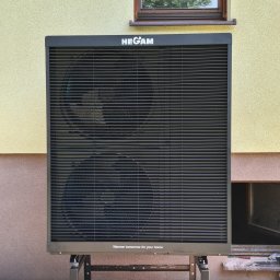 HEGAM - Czarna pompa ciepła HEGAM zamontowana na zewnątrz budynku z oknem, z widocznymi dwoma wentylatorami i napisem 'Warmer tomorrow for your home'.