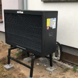 Czarna pompa ciepła marki HEGAM zamontowana na metalowym stelażu z betonowymi podstawami, widoczna na tle elewacji budynku i okna z brązowymi okiennicami, teren wokół w trakcie prac ziemnych.