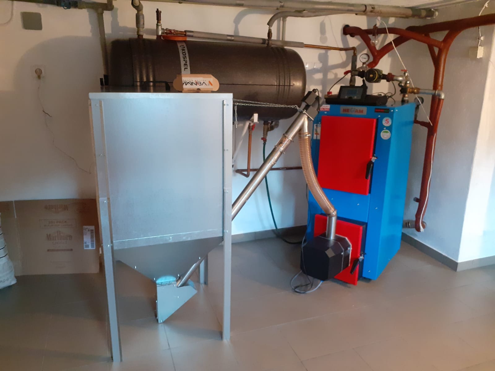 Kotłownia z piecem na paliwo stałe z podajnikiem, zbiornikiem buforowym i instalacją hydrauliczną w piwnicy, widoczne kartony na podłodze.