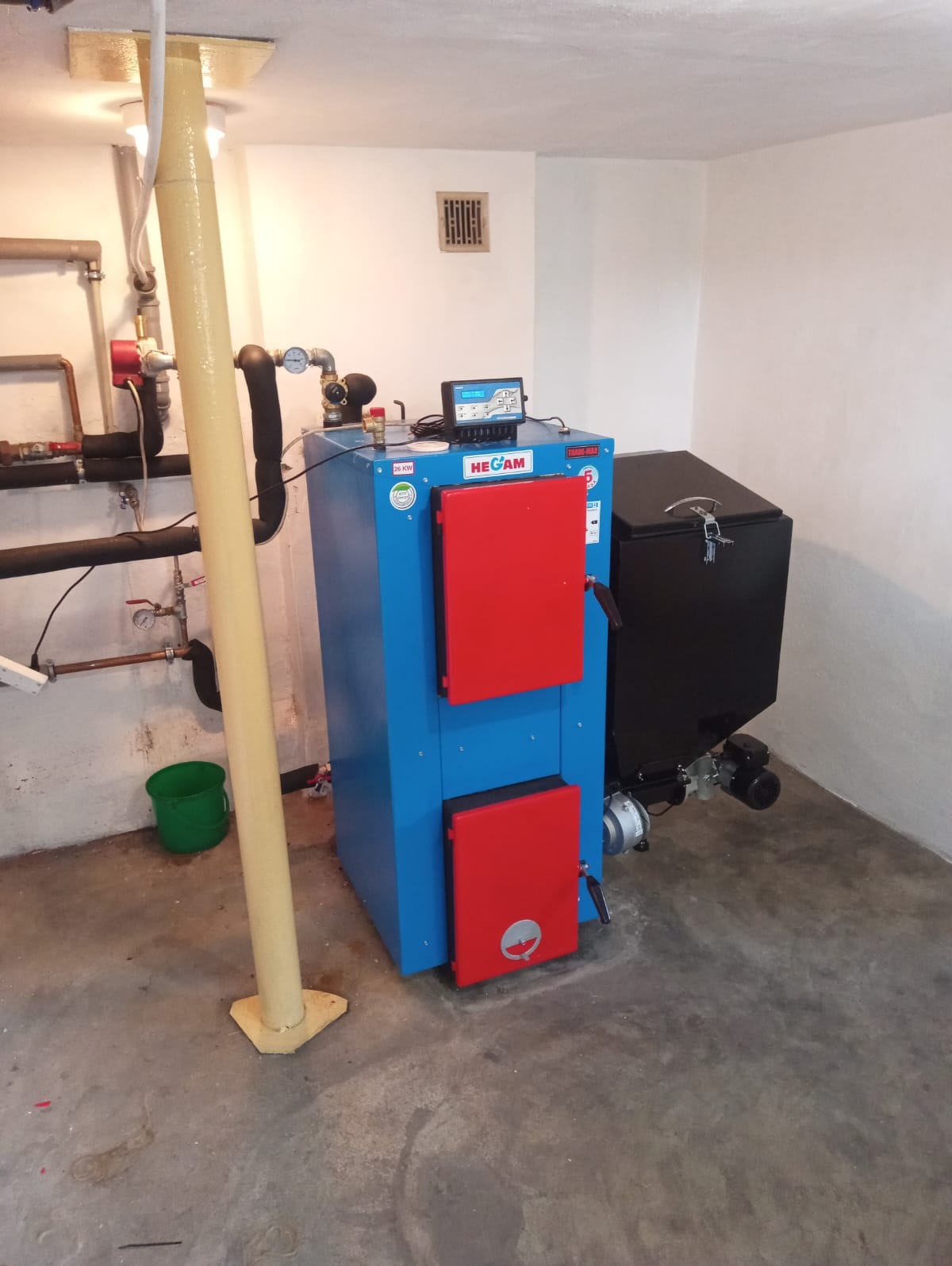 Niebieski piec na paliwo stałe z czerwonymi drzwiczkami marki HECAM, z czarnym podajnikiem, zamontowany w kotłowni z widocznymi rurami i zielonym wiadrem.