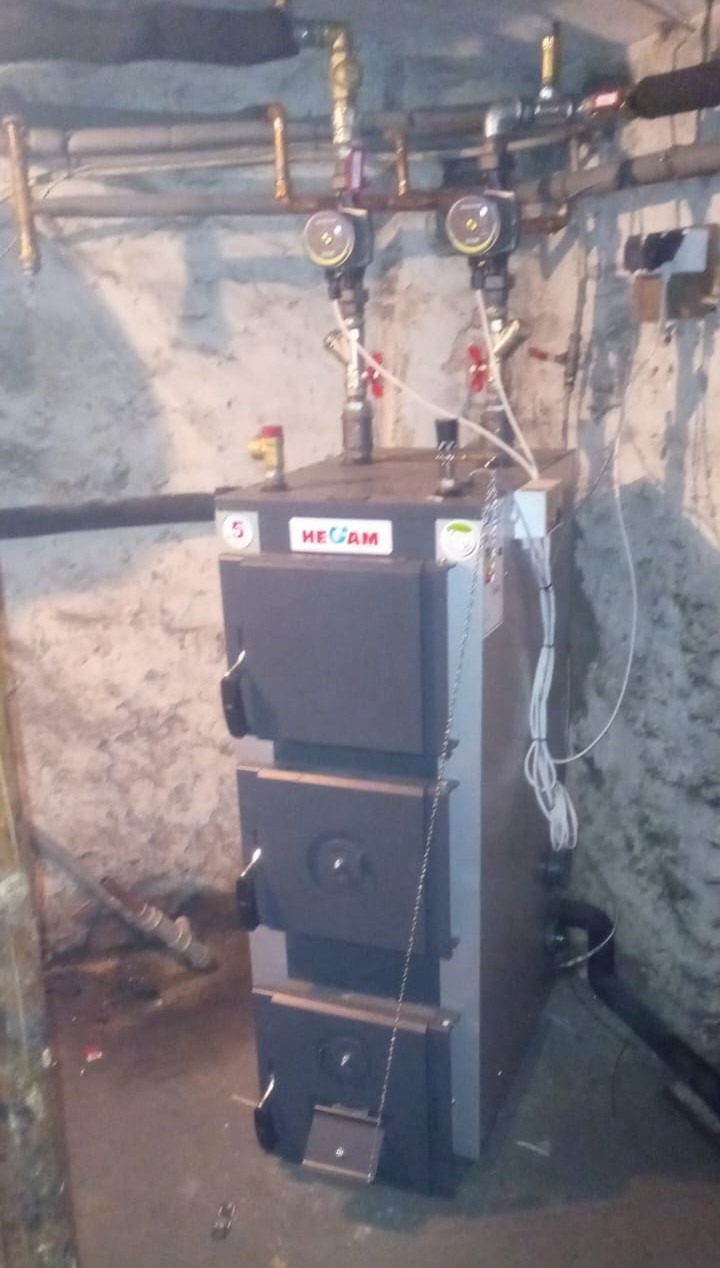 Nowy, trzykomorowy piec na paliwo stałe marki HEGAM, podłączony do instalacji grzewczej z dwoma pompami obiegowymi, zamontowany w piwnicy o surowych, betonowych ścianach.