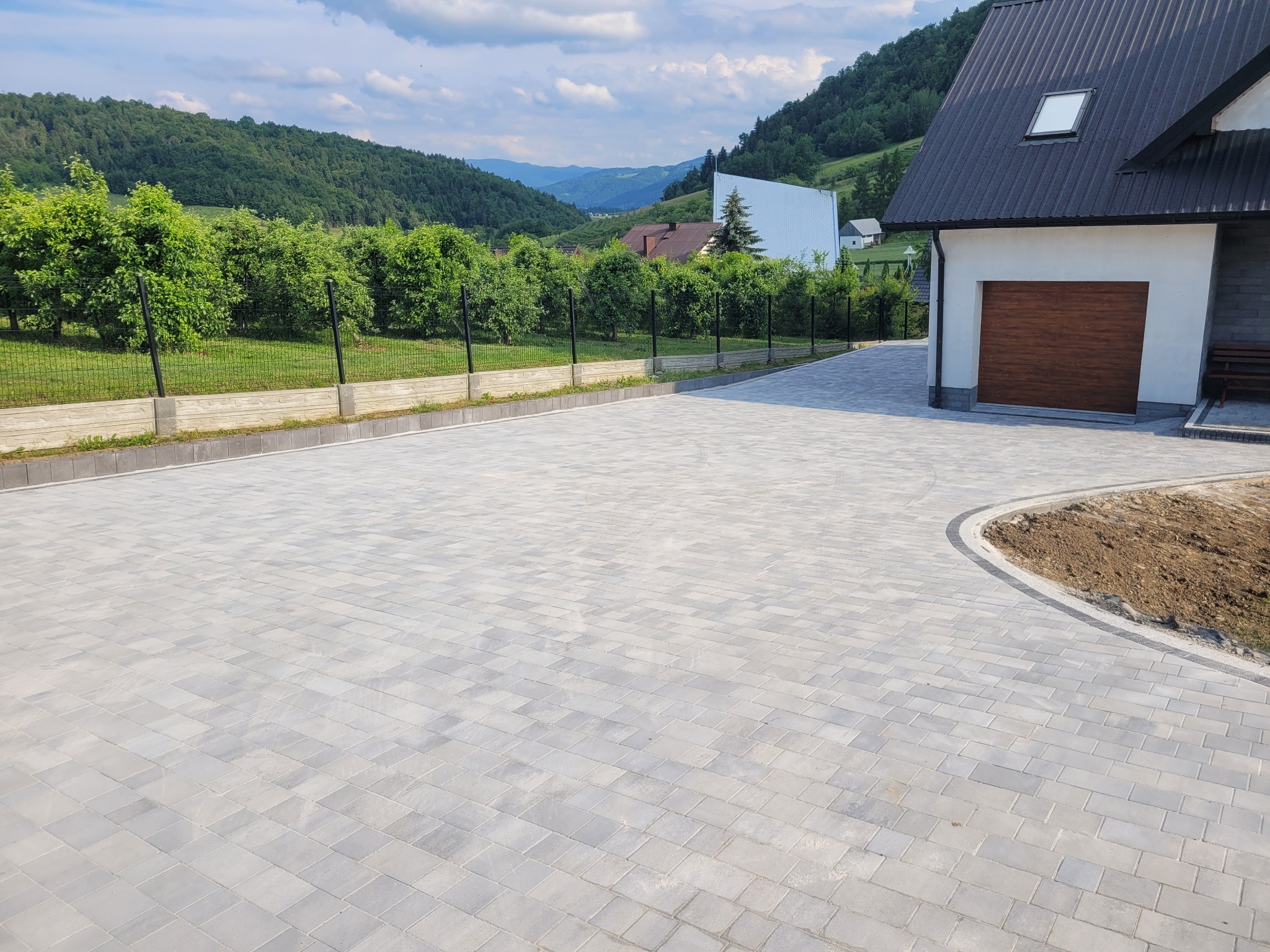 Nowo ułożona, szara kostka brukowa na podjeździe domu z brązową bramą garażową, otoczona zielenią i górskim krajobrazem w tle.