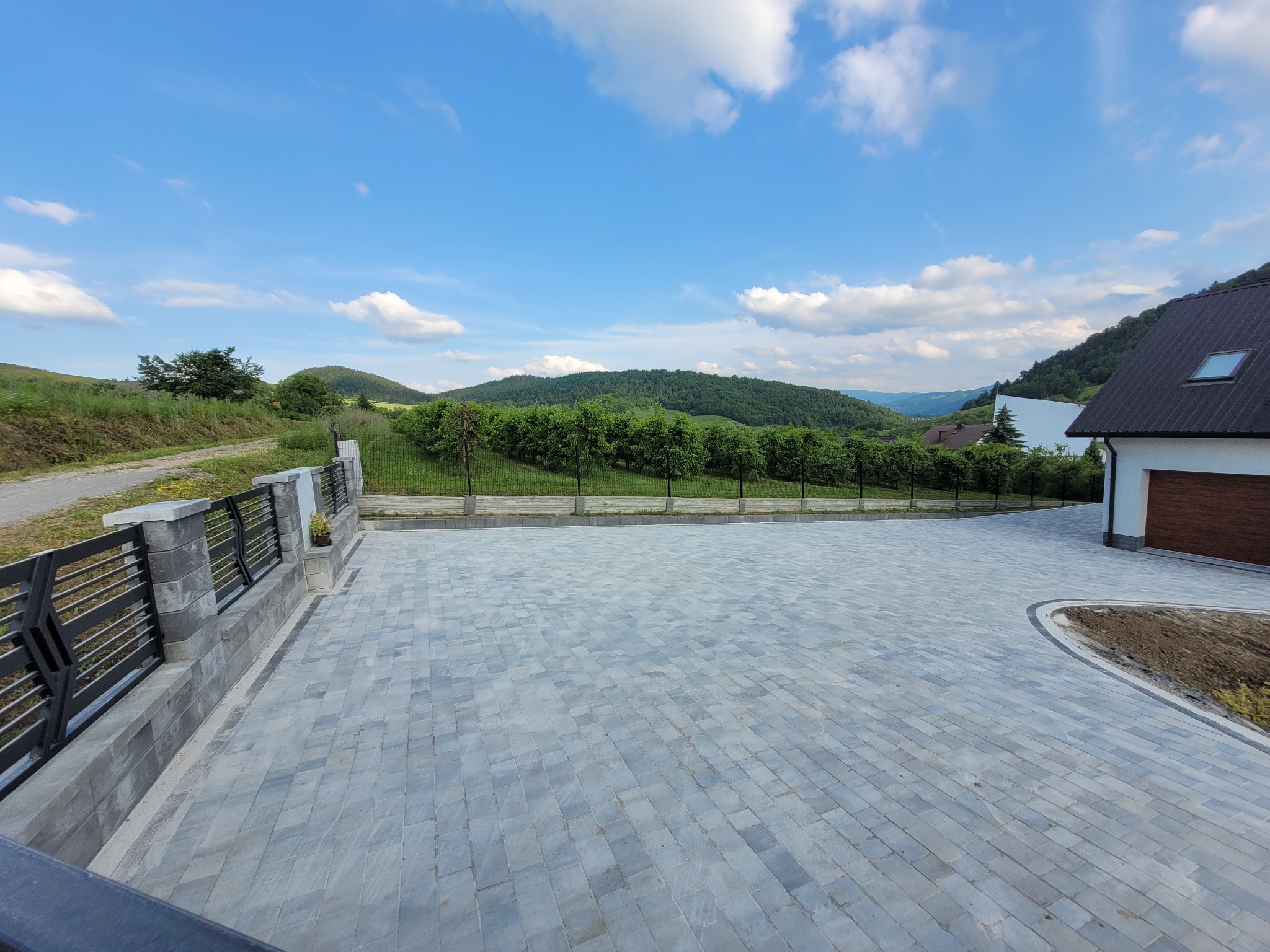 Szara kostka brukowa ułożona na posesji w Łącku, z widokiem na wzgórza i błękitne niebo z chmurami, metalowe ogrodzenie w tle.