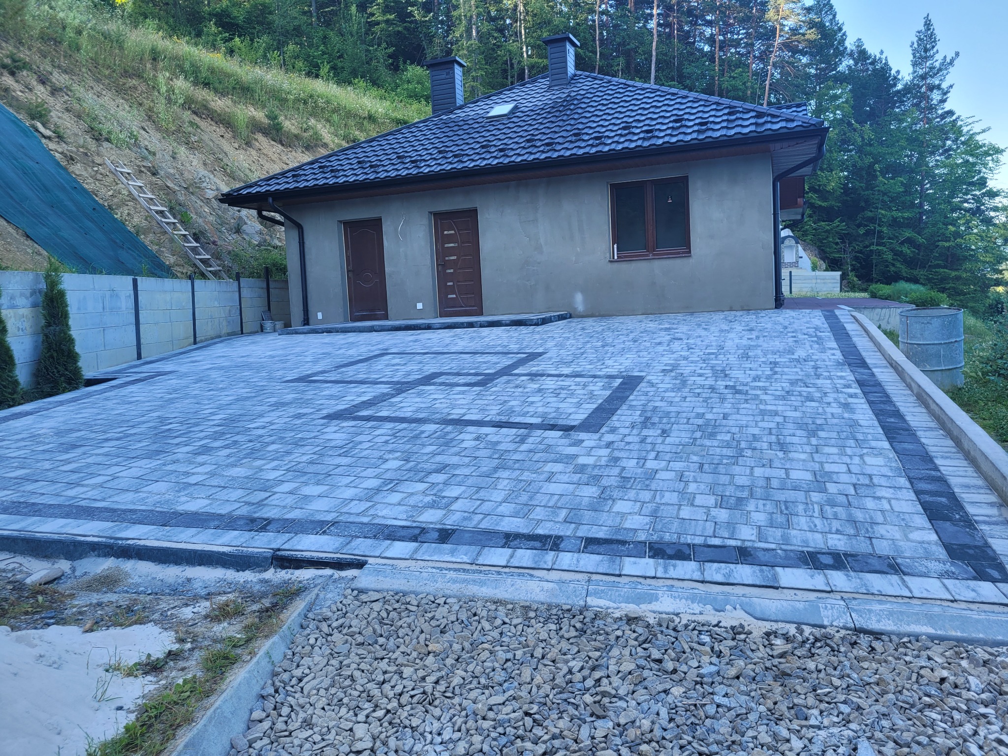 Ułożona kostka brukowa w geometryczny wzór przed nowo wybudowanym domem z szarą elewacją i ciemną dachówką, widok na zbocze porośnięte drzewami w tle.