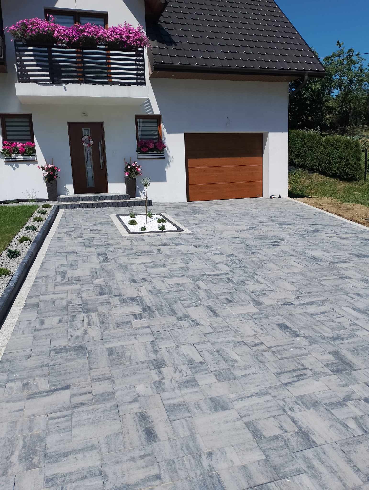 Ułożona szara kostka brukowa przed domem z białymi ścianami, brązowymi drzwiami i bramą garażową, rabatami z białym żwirem i zielenią.