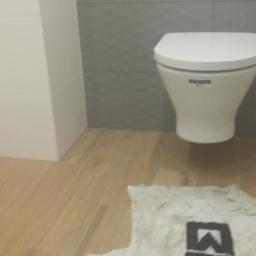 Firma remontowo budowlana Robert lech - Nowoczesna toaleta wisząca na tle szarej, strukturalnej ściany, z drewnopodobną podłogą i roboczą szmatką z logo na pierwszym planie.