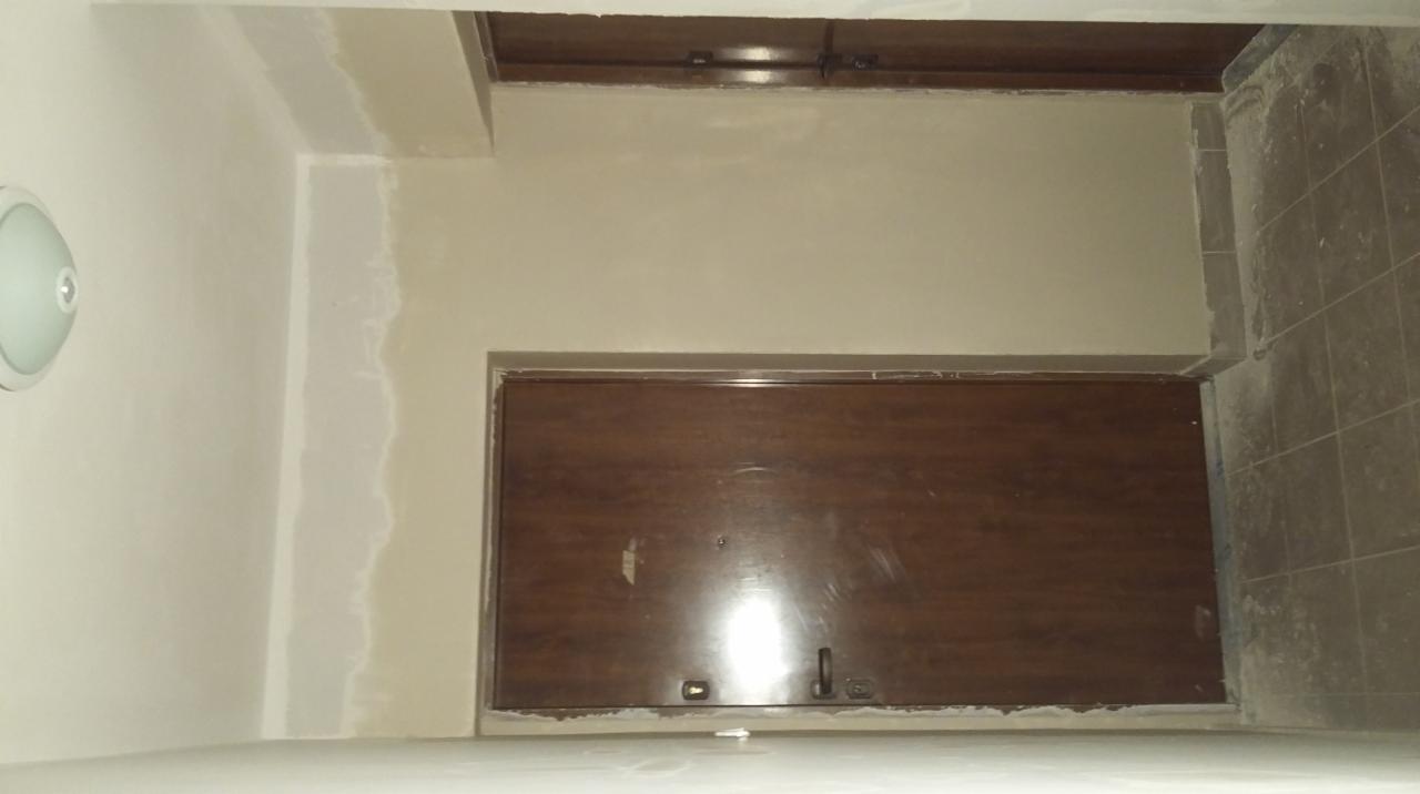 Wnętrze pomieszczenia w trakcie prac wykończeniowych: ściany częściowo wyszpachlowane, widoczna zabudowa szafkowa i fragment ułożonej, szarej glazury podłogowej.