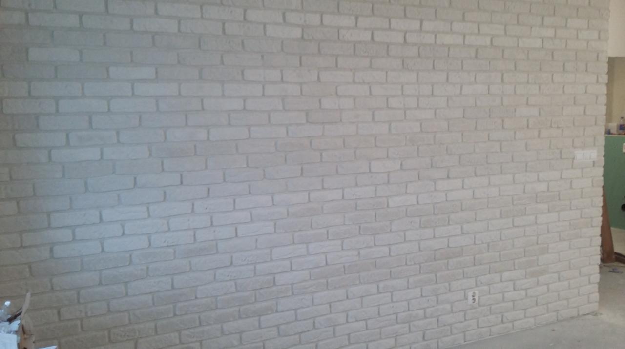 Ściana wykończona białymi cegłami dekoracyjnymi, widoczne gniazdko elektryczne i włącznik światła, fragment zielonej ściany w tle.