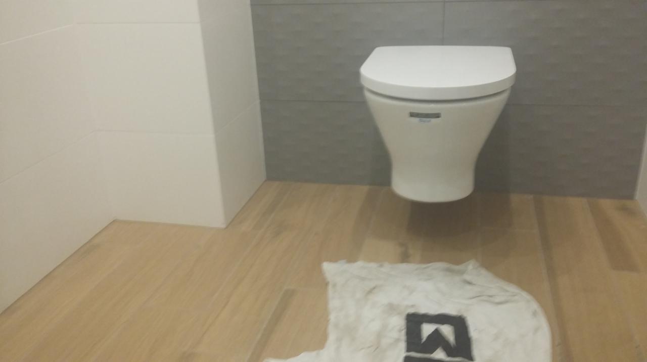 Nowoczesna toaleta wisząca na tle szarej, strukturalnej ściany, z drewnopodobną podłogą i roboczą szmatką z logo na pierwszym planie.