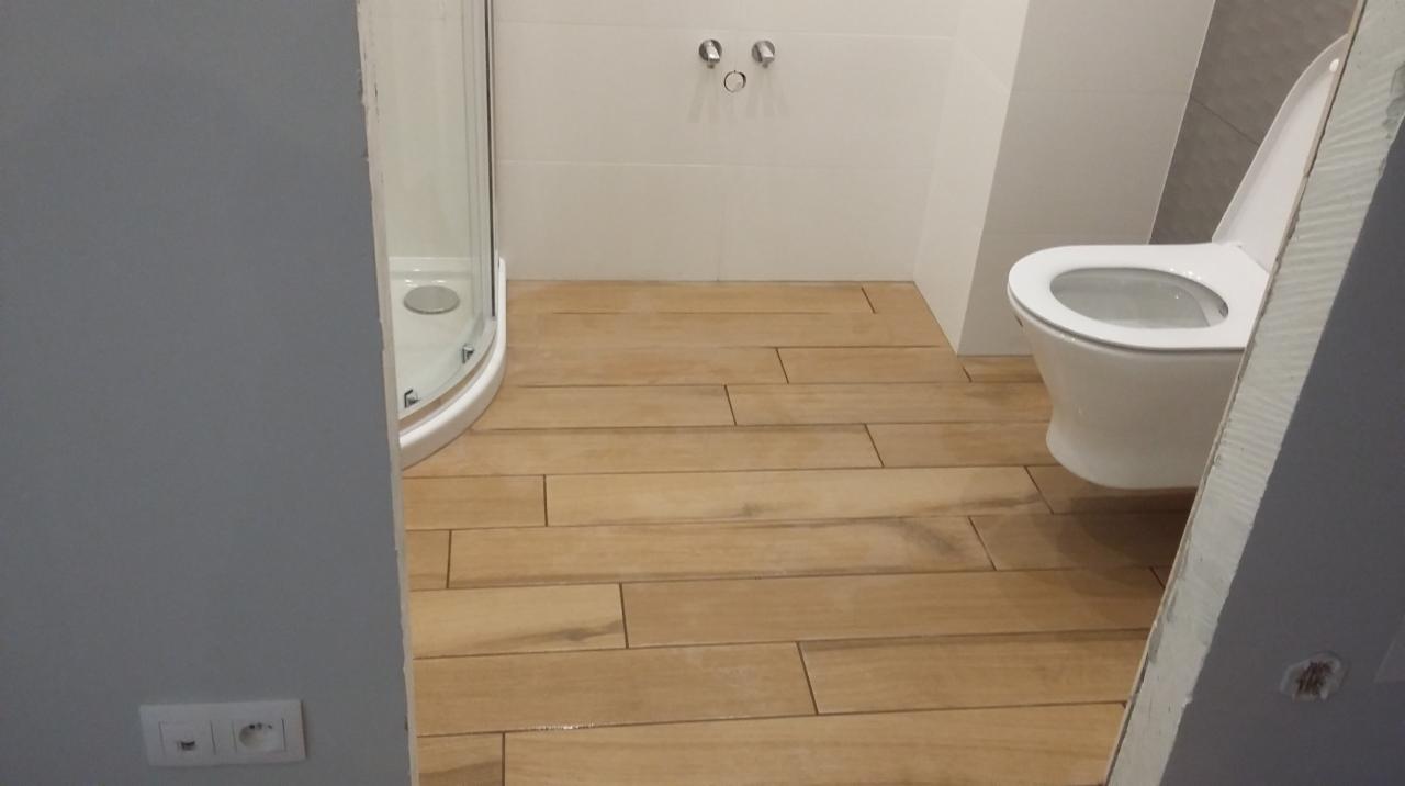 Wykończona łazienka z kabiną prysznicową, toaletą i płytkami podłogowymi imitującymi drewno ułożonymi w cegiełkę.