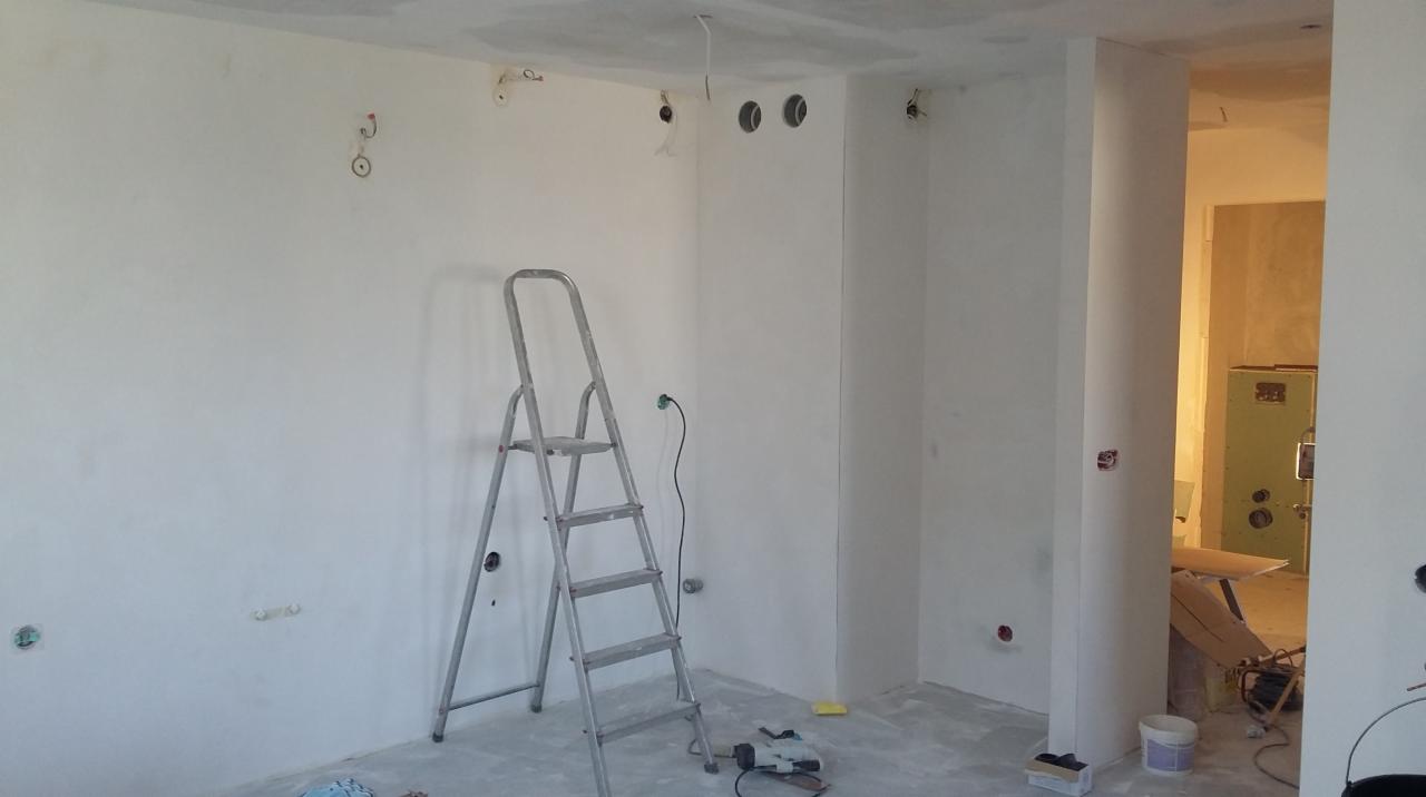 Pokój w trakcie prac wykończeniowych: ściany po szpachlowaniu, widoczne otwory na gniazdka, drabina aluminiowa, szlifierka na podłodze, surowy beton na suficie.