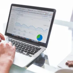 Ręce mężczyzny piszącego na laptopie z wykresem analitycznym na ekranie, obok talerz z ciasteczkami w kształcie gwiazdek.