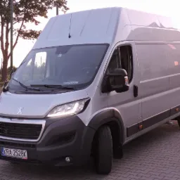 Srebrny bus marki Peugeot z włączonymi światłami postojowy, zaparkowany na kostce brukowej przed ogrodzeniem z siatki.